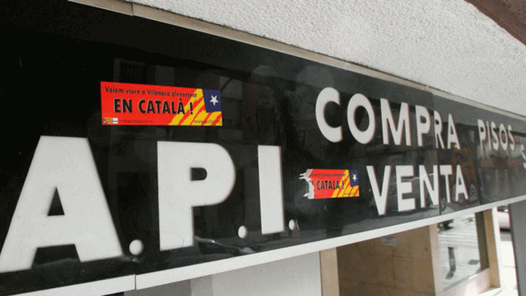 Rótulo de una inmobiliaria en castellano, con una pegatina que exige el catalán