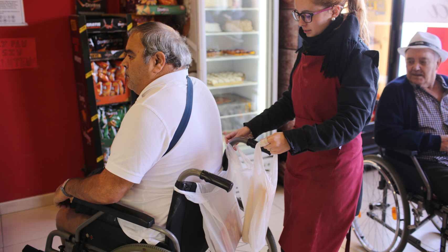 Una dependienta de panadería cuelga en la silla de ruedas de José Bernal dos barras de pan y un paquete de chorizo que acaba de comprar.