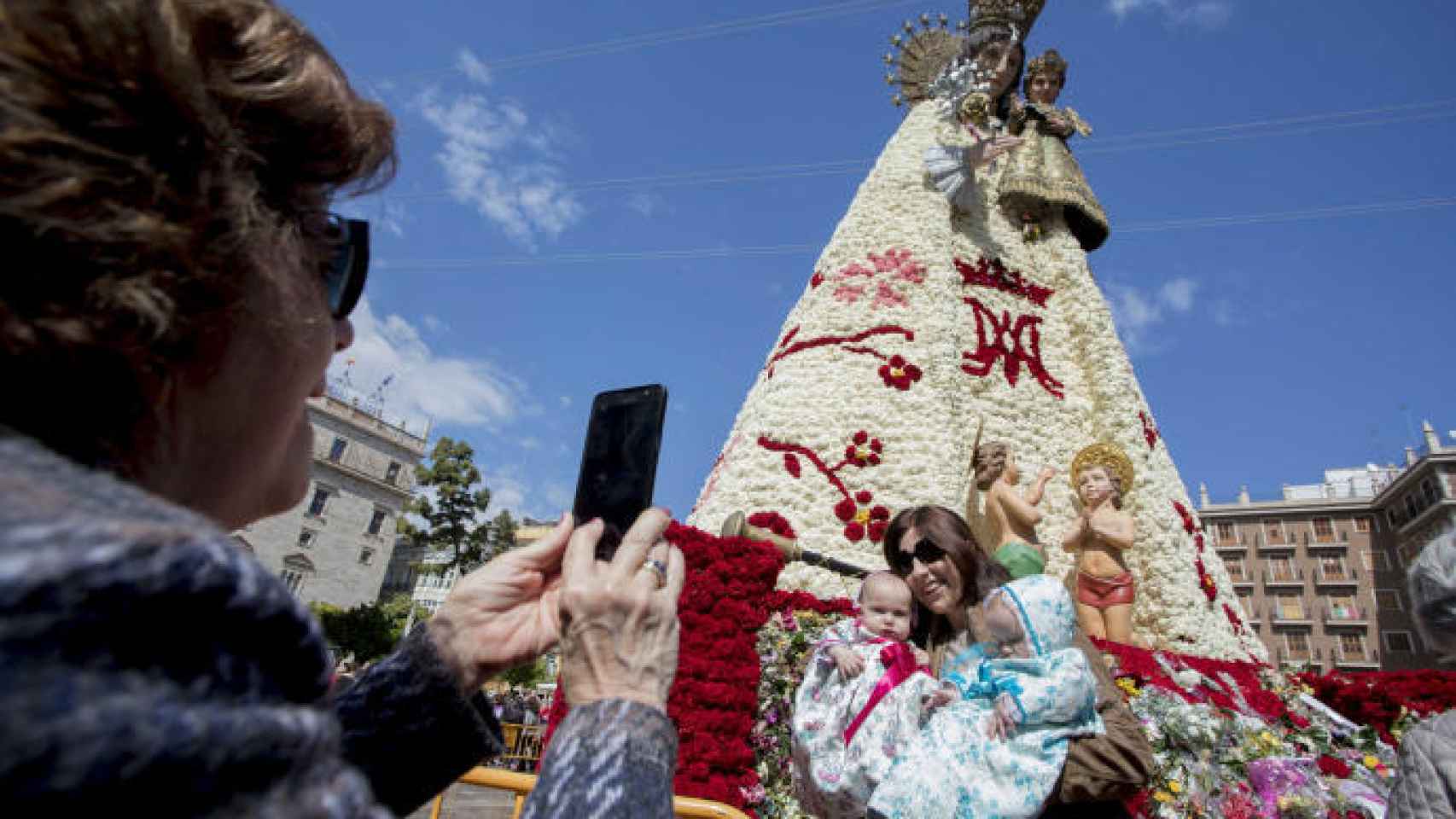 Una señora hace una foto a una joven con dos menores delante del manto de la Virgen cubierto de ramos de flores tras la ofrenda.