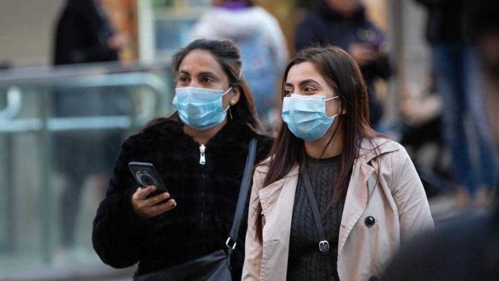 Dos mujeres pasean por la calle con mascarillas en la cara y consultando su teléfono móvil.