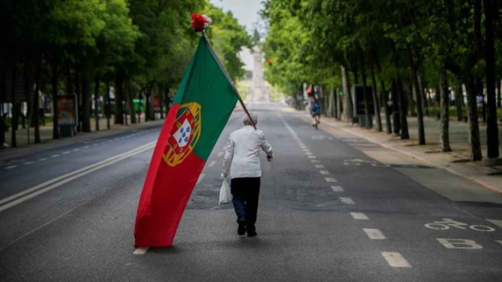 Un ciudadano porta una bandera portuguesa en una avenida solitaria.