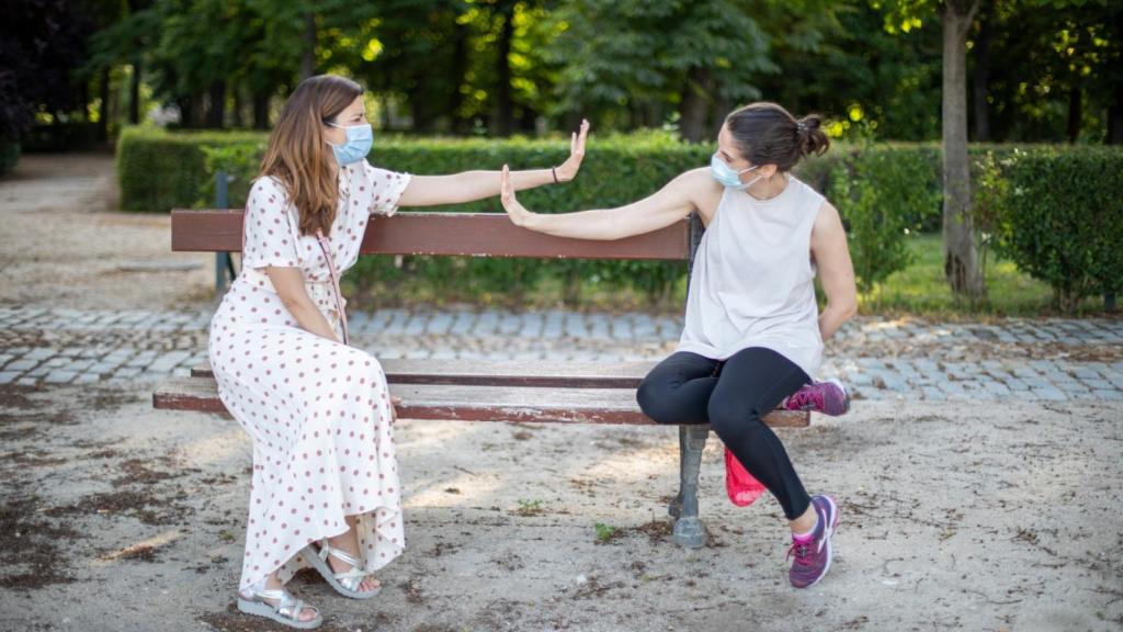 Dos amigas simulan saludarse sentadas en un banco del céntrico parque madrileño.