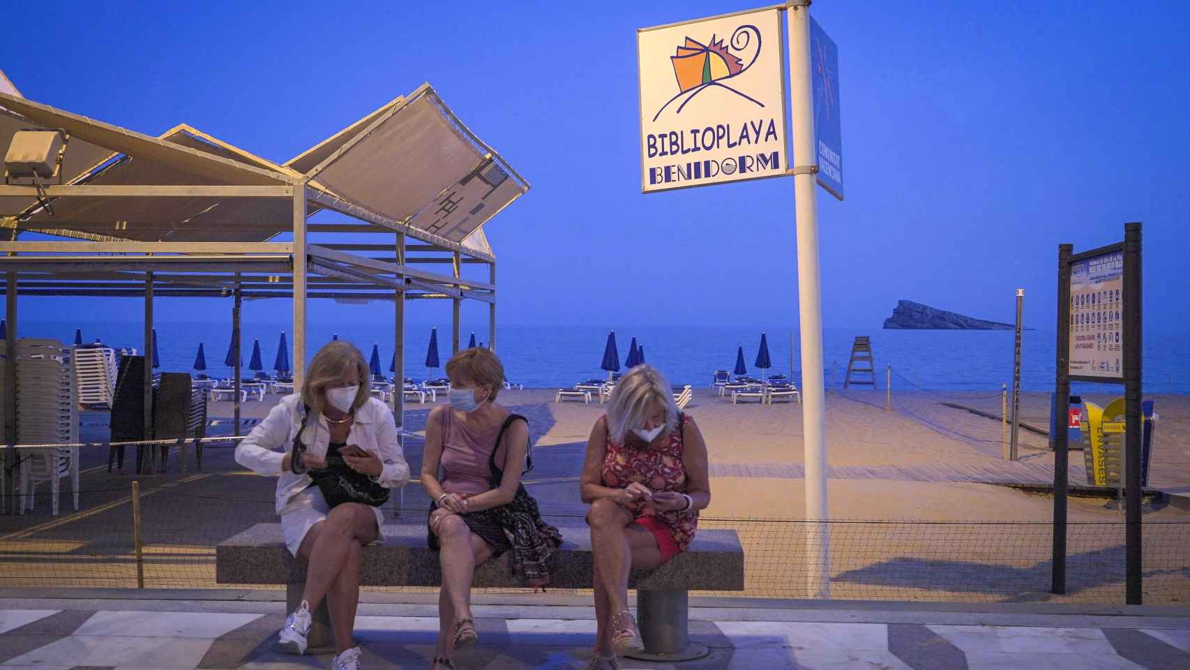 Tres personas con mascarillas sentadas frente a la playa de Benidorm.