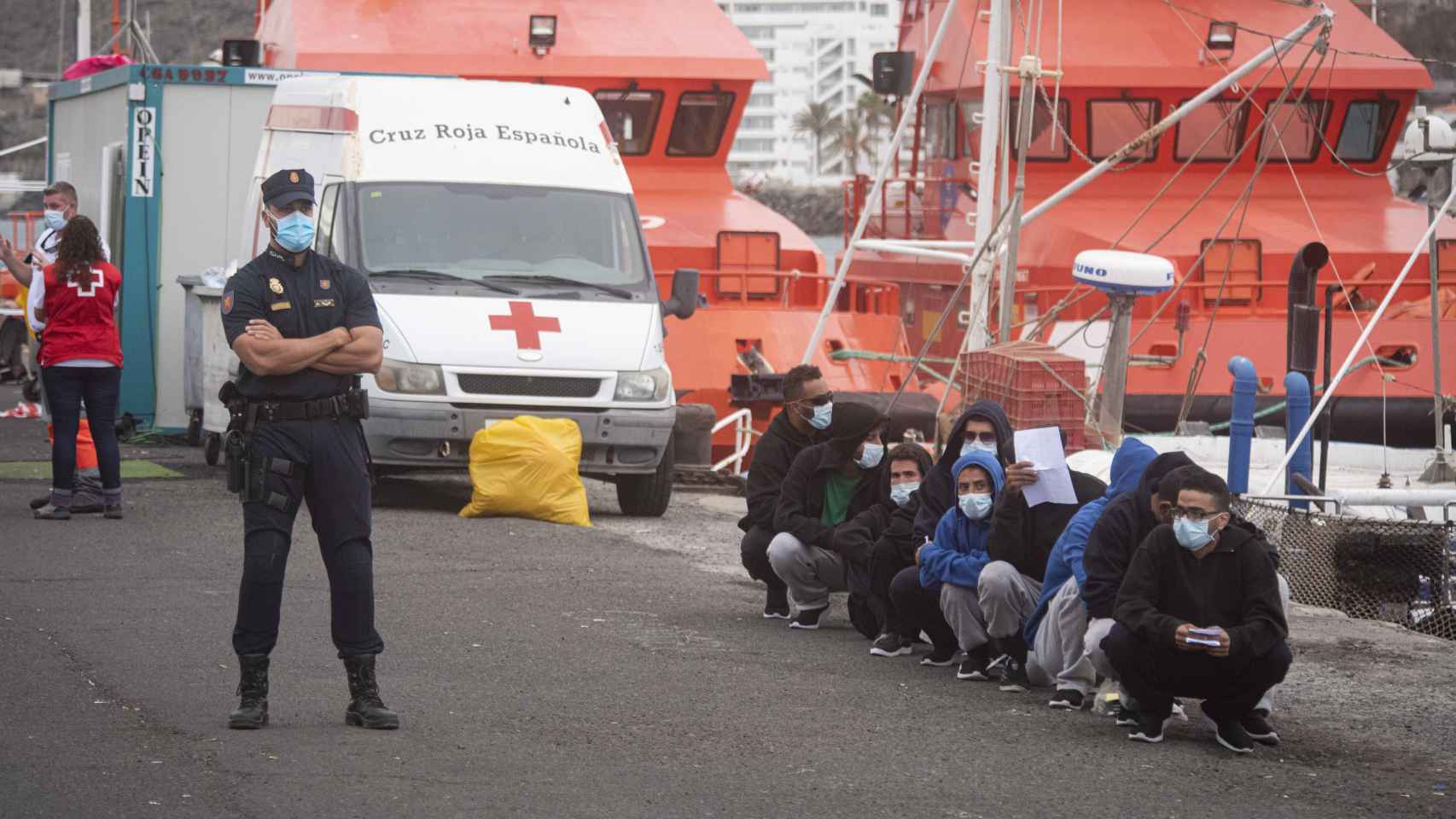 Un policía custodia a un grupo de inmigrantes que espera su traslado fuera del muelle de Arguineguín.