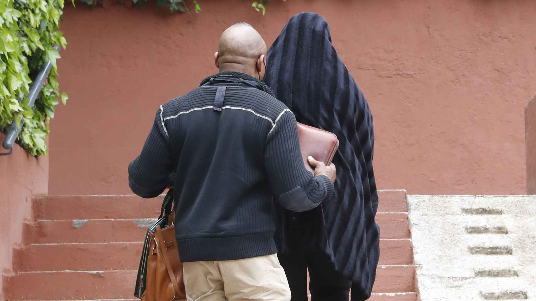 Raquel llegando a su casa oculta bajo una manta negra y ayudada por su marido, Isi.
