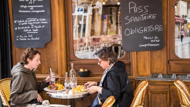 Dos clientas almuerzan en una terraza de París bajo un letrero donde se lee pase sanitario obligatorio.