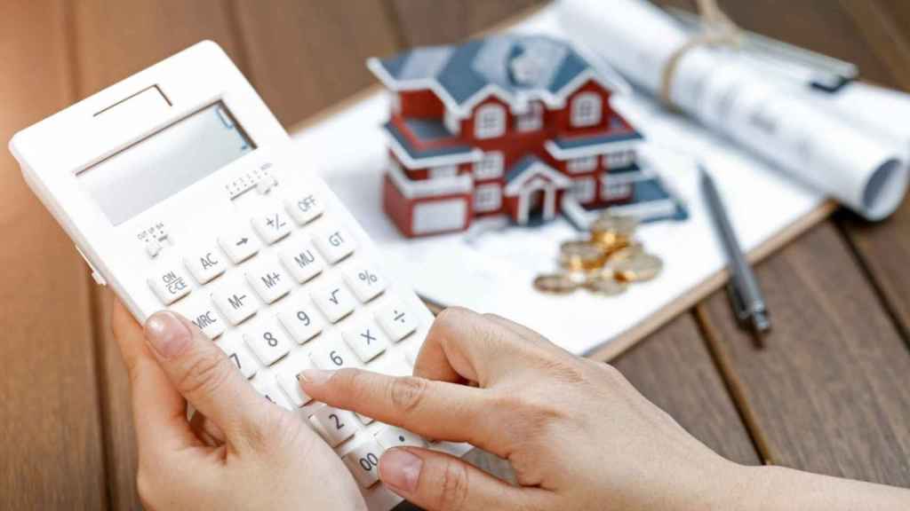 Imagen de una persona usando una calculadora delante de la maqueta de una casa.