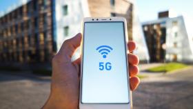 Una mano sujeta un smartphone con el logo del 5G en la pantalla delante de unos edificios.