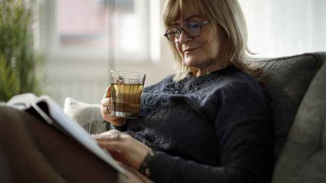 Mujer madura sonriente leyendo un libro y bebiendo té en casa