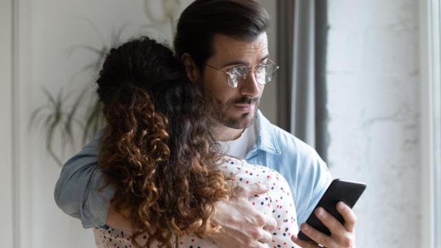 Hombre abrazando a una mujer y revisando su teléfono. Foto: iStock.